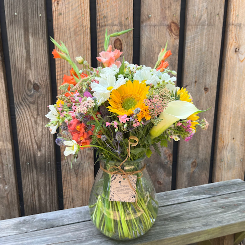A large, colorful bouquet of seasonal blooms arranged in a clear glass vase, placed on a wooden surface, with a tag attached that reads 'Good Karma Bouquet'.